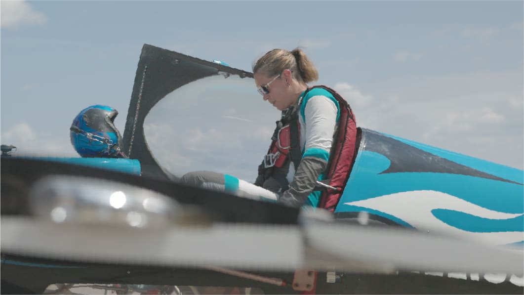 A pilot climbing into the cockpit of a teal and white race aircraft on the ground, helmet resting on the nose