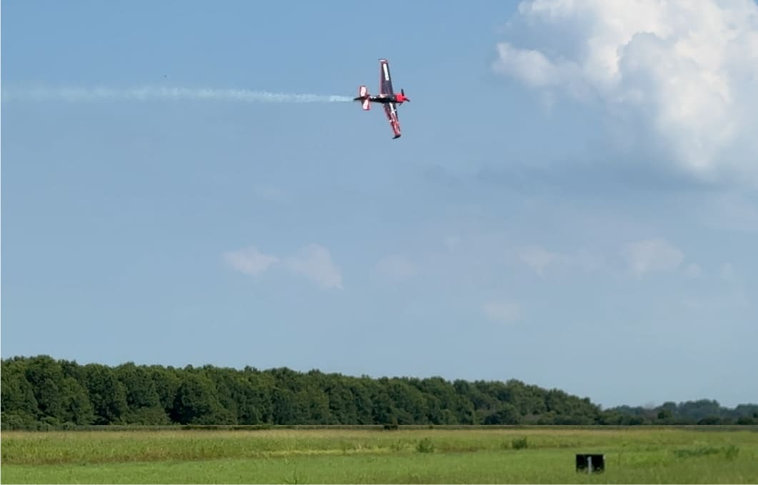 A red and white race aircraft banked hard with a smoke trail streaming behind it over a green field