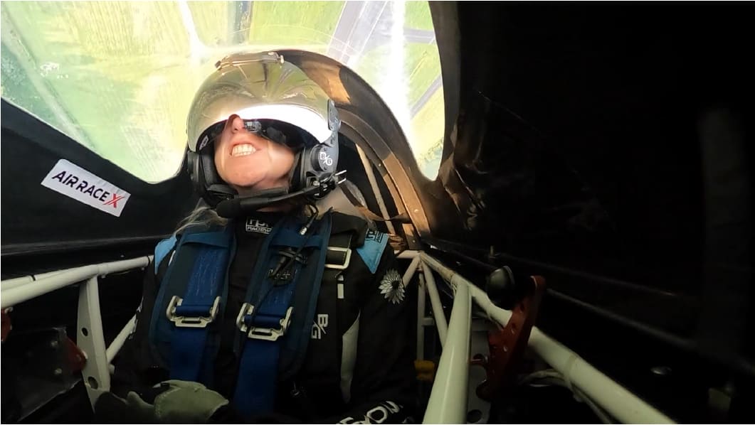 Cockpit view of a smiling pilot pressed back into her seat during a high-G turn, with the ground visible through the canopy overhead