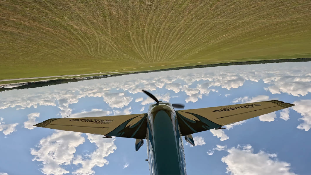 Wing-mounted camera view of an aircraft flying inverted low over the airfield, clouds below and ground above